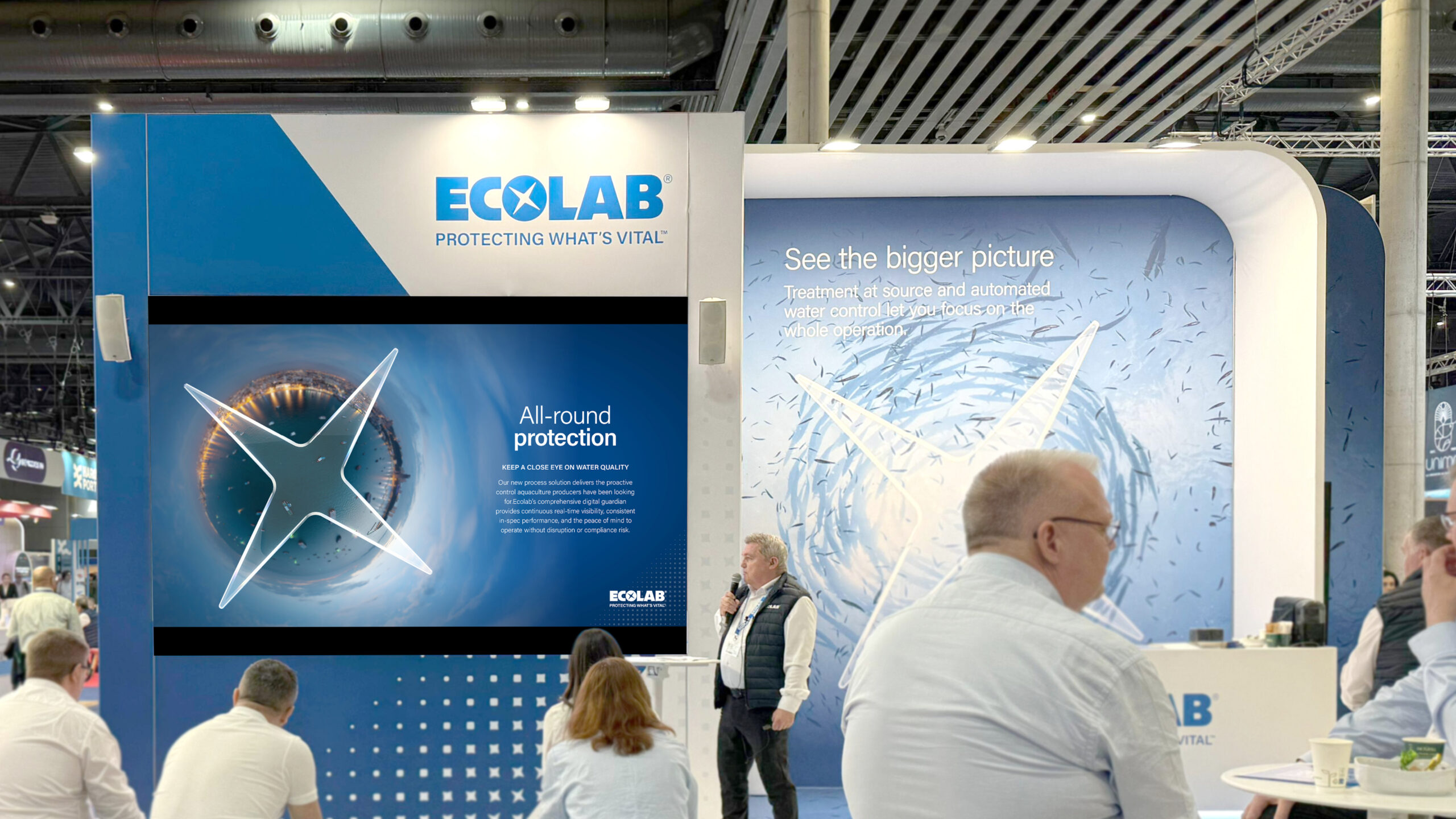 A presenter speaking to an audience at an Ecolab exhibition booth with large branded displays reading 'All-round protection' and 'See the bigger picture'
