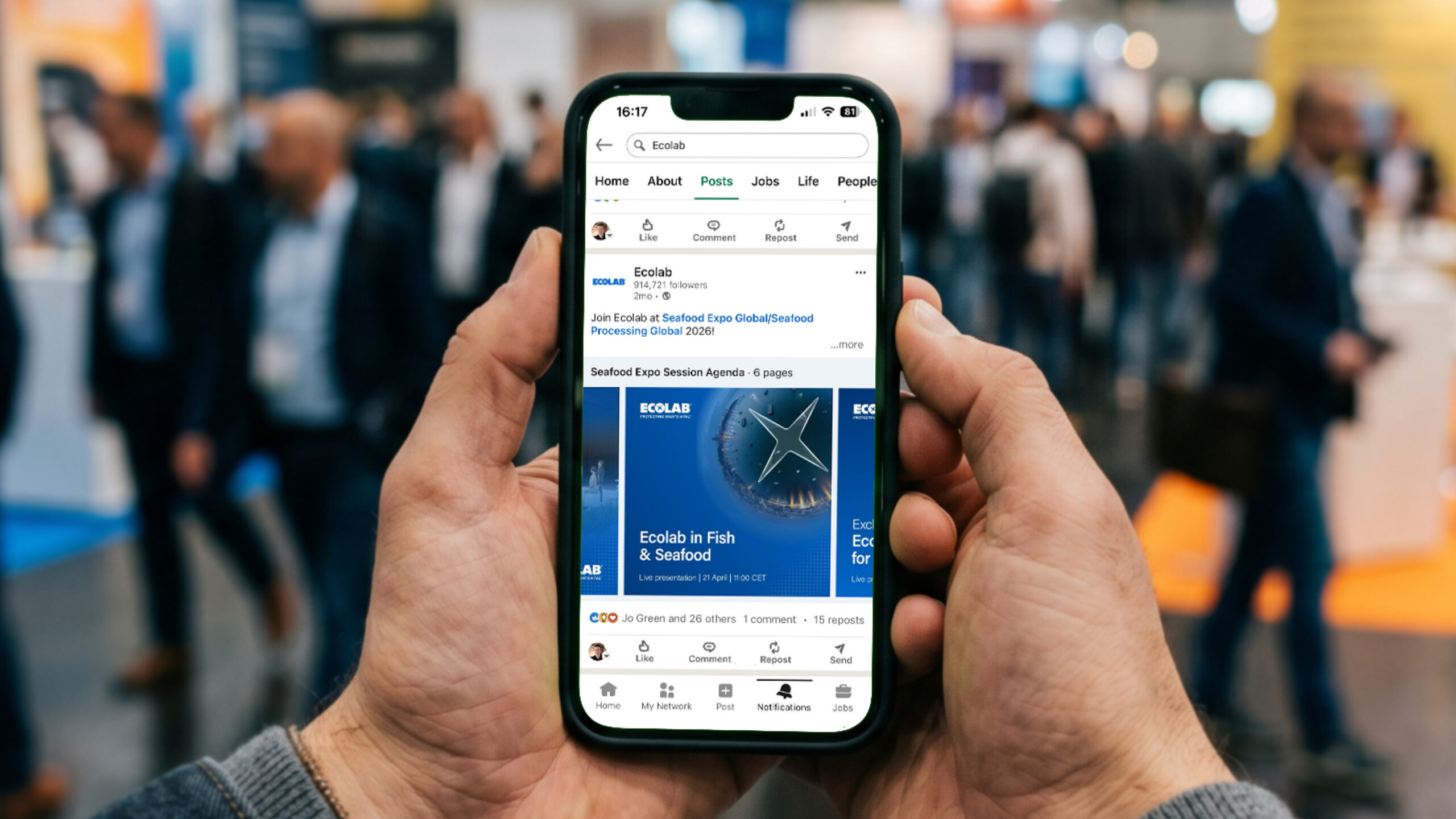 Hands holding a smartphone in a busy expo hall, displaying an Ecolab LinkedIn post promoting their 'Ecolab in Fish & Seafood' presentation at Seafood Expo Global.